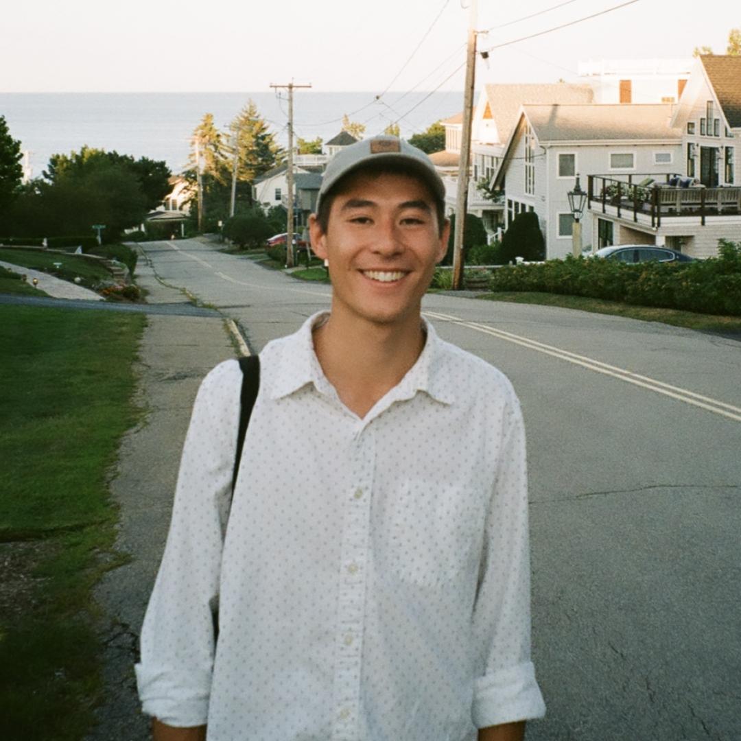 [Image Description: Commonwealth Corps Intern, Matt Lee, an Asian male wearing a gray hat and white dotted button up shirt. He is smiling with a street and houses in the background.]
